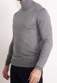 Mężczyzna w dopasowanym szarym swetrze z golfem i ciemnoniebieskich dżinsach, stojący na tle jasnego, jednolitego tła.