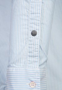 Ljusblå randig skjorta med en slät textur, med en svart mönstrad knapp och en knapp med rosa detaljer på manschetten.