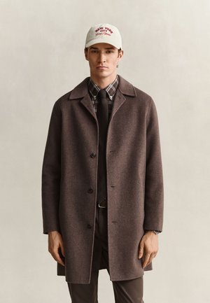 Homme portant un manteau marron boutonné, une chemise à carreaux, une cravate noire, un pantalon marron et une casquette blanche avec un texte rouge "New York City" sur un fond uni.