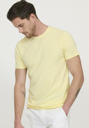 T-shirt en coton jaune à manches courtes, col rond et coupe slim. Présente une texture lisse et un design minimaliste, assortie à un pantalon blanc.