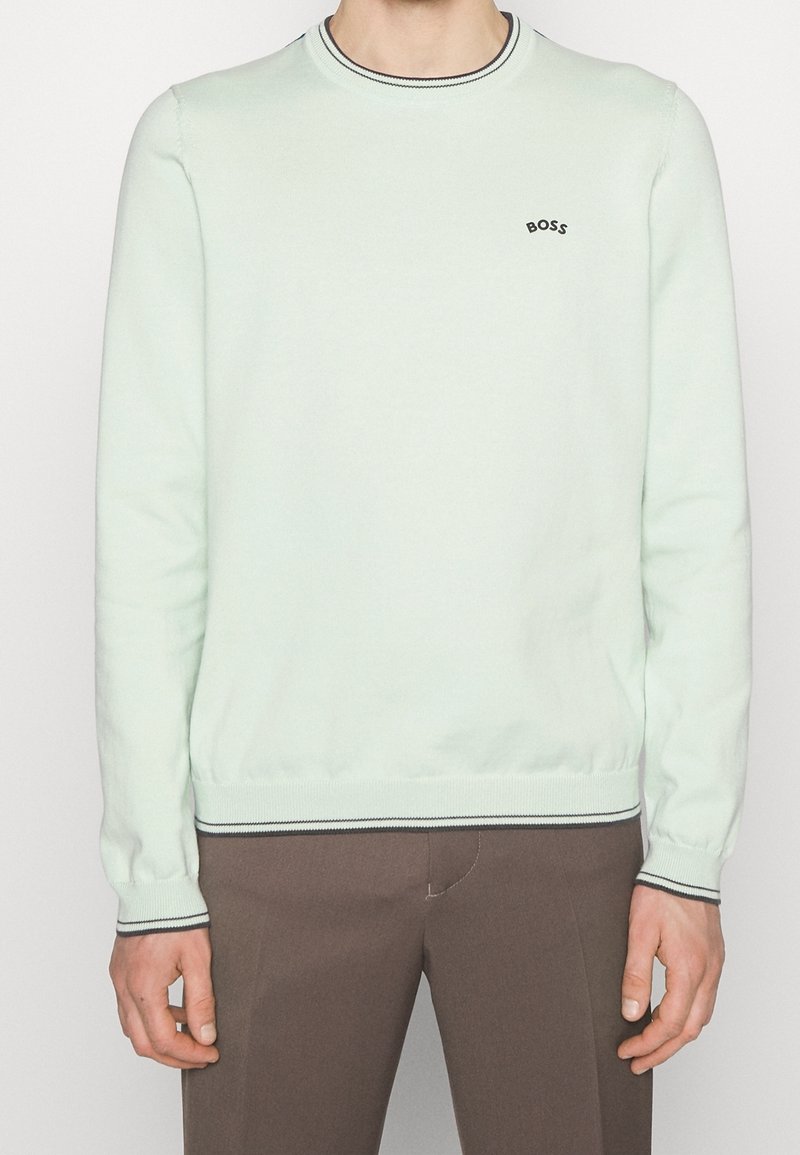 Homme portant un pull à manches longues vert menthe clair avec des bordures noires et un petit logo "BOSS" sur la poitrine, associé à un pantalon marron.