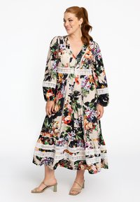 Florales Maxikleid mit mehrfarbigen Drucken auf einem cremigen Hintergrund, langen Ärmeln, einem V-Ausschnitt und Spitzenbesatz. Kombiniert mit grünen Absatzsandalen.