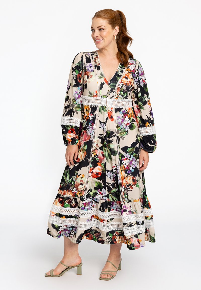 Florales Maxikleid mit mehrfarbigen Drucken auf einem cremigen Hintergrund, langen Ärmeln, einem V-Ausschnitt und Spitzenbesatz. Kombiniert mit grünen Absatzsandalen.