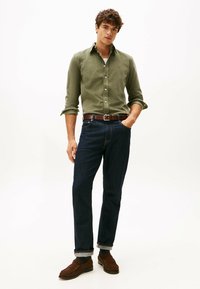 Camisa verde oliva de botones, pantalones vaqueros azul oscuro con los puños remangados, cinturón de cuero marrón y mocasines con calcetines oscuros, accesorios mínimos.