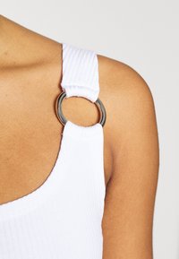 Primer plano de un hombro que lleva una blusa de tela acanalada blanca con un detalle de anillo plateado que conecta la tira.