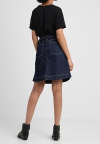 Corte A en falda de mezclilla con un lavado azul oscuro, costuras visibles, y dos bolsillos traseros. Combinada con una camiseta negra y botas de tobillo.