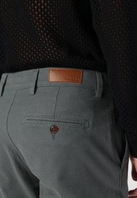 Pantaloni grigi chiari con finitura testurizzata, una toppa di pelle con marchio e una tasca posteriore con chiusura a bottone.