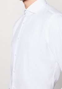 Biała koszula z guzikami i usztywnionym kołnierzykiem, wykonana z teksturowanej tkaniny z delikatnym połyskiem. Sześć guzików z przodu.