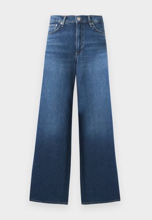 Kvinde stående iført en hvid ærmeløs top, blå vidde jeans med sort bælte og brune sandaler mod en ensfarvet hvid baggrund.