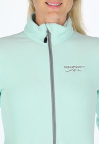 Light turquoise fleece jacket with a high collar and gray zipper. Features 'SWEDEMOUNT' logo in gray on the front. Soft texture.
