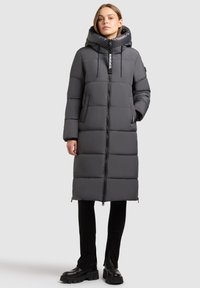 Ženska v dolgem, sivem prešitem plašču s kapuco, črnih hlačah in debelih črnih škornjih, stoji ob svetlo sivi ozadju.