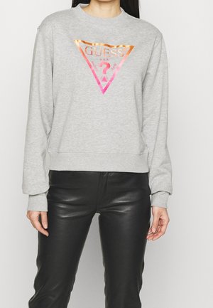 Femme portant un sweat-shirt GUESS gris clair avec un logo triangle brillant et un pantalon noir en similicuir, debout devant un fond blanc.