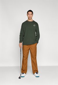 Chemise à manches longues vert foncé avec logo, pantalon beige et chaussures de golf blanches, tenant un club de golf. Tenue de golf simple et décontractée.