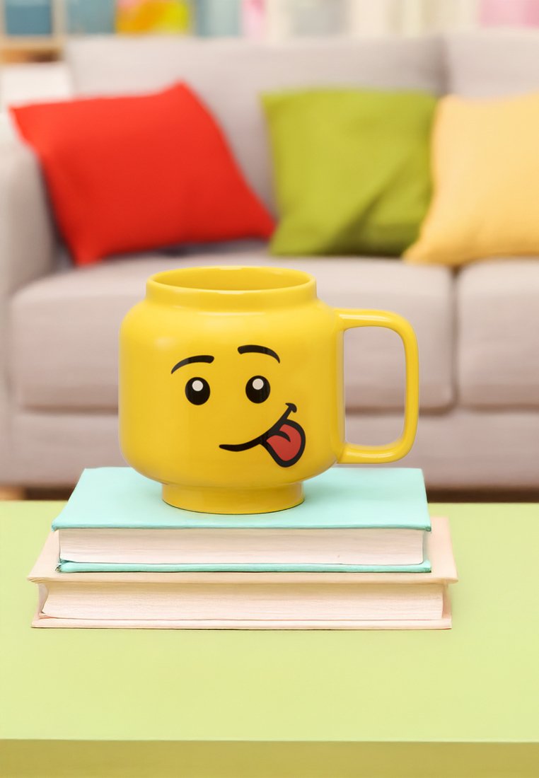 Mug en céramique jaune en forme d'emoji souriant, affichant une expression avec la langue tirée, posé sur des livres empilés avec une table verte en dessous.