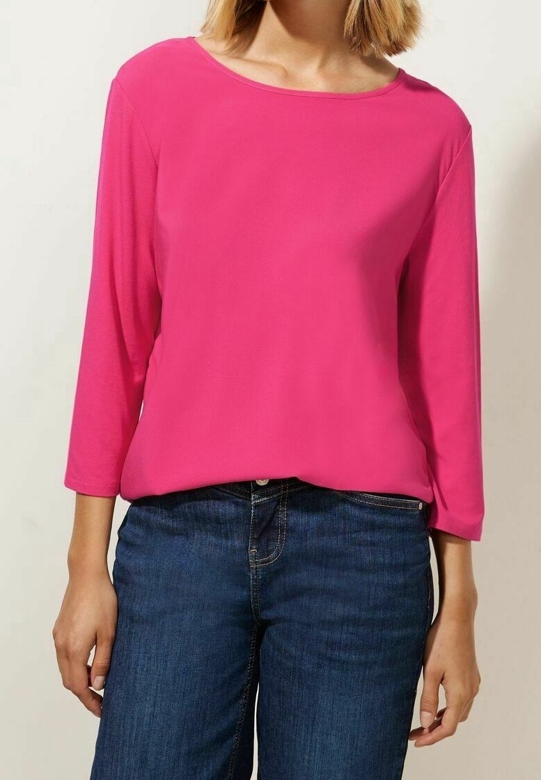 Femme portant un haut rose vif à manches longues avec un col rond et un jean bleu, debout devant un fond uni.