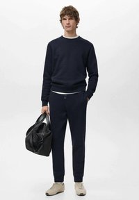 Marineblauwe trui en broek van een zacht materiaal, gecombineerd met beige sneakers. Zwarte sporttas vastgehouden in één hand, met handvatten en ritssluiting.