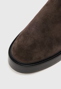 Bottines en suede marron, présentant un bout arrondi et une épaisse semelle en caoutchouc noir. Texture lisse avec un léger éclat sur la surface supérieure.