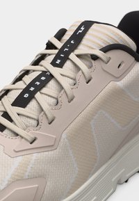 Κοντινό πλάνο μπεζ αθλητικού παπουτσιού με σταυρωτά κορδόνια και μαύρη λωρίδα με λευκό κείμενο στη γλώσσα.