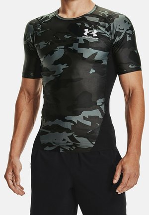Mand iført tætsiddende sort og grå camouflage Under Armour kompressions-T-shirt med korte ærmer og sorte shorts, stående med armene afslappet ned langs siden.
