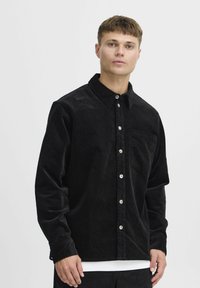 Zwarte corduroy button-up shirt met een kraag, een borstzak en metalen knopen. De stof heeft een zachte, gestructureerde afwerking.