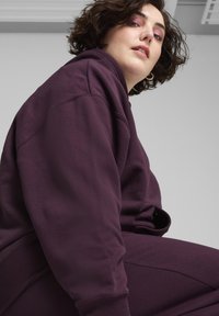 Felpa color prugna e pantaloni abbinati in materiale morbido. Presenta polsini e orlo a costine. Il modello posa con un leggero angolo verso l'alto, rivelando la texture del tessuto.