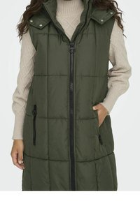 Woman wearing a long olive green quilted vest with front zipper and zippered pockets over a beige cable-knit sweater.