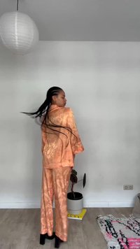 Femme aux longs cheveux tressés portant un costume orange en soie qui balance ses cheveux dans une pièce blanche avec une plante, un tapis, une lampe en papier suspendue et un sol en bois clair.