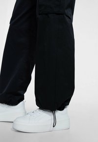 Jambes portant un pantalon noir avec des cordons ajustables aux chevilles et des baskets blanches à plateforme avec lacets sur fond neutre.