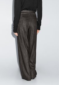 Persoon in een hoge taille, brede broek met donkergrijze streepjes en een zwart lange mouwen top, staande met de handen ontspannen langs de zij.