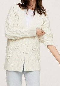 Woman wearing a white cable-knit cardigan with floral accents, buttoned front, and rolled-up sleeves over a white shirt and light jeans.
