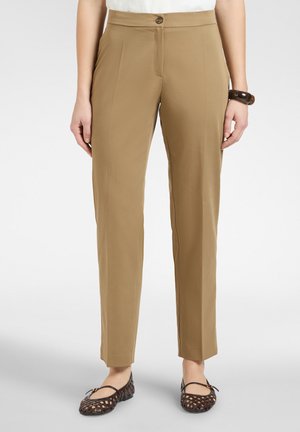 Frau trägt hellbraune gerade geschnittene Hosen, ein weißes, hineingestecktes Oberteil, dunkelbraune gewebte Ballerinas und ein dunkles Armband am rechten Handgelenk.