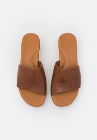 Une paire de sandales ouvertes en cuir marron à enfiler, avec de larges sangles en cuir et des semelles intérieures rembourrées portant l'inscription "SUPER SOFT", sur fond blanc.