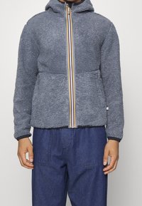Giacca in pile di colore grigio scuro con finitura testurizzata, dotata di una zip multicolore, cappuccio e polsini elastici, abbinata a pantaloni in denim.