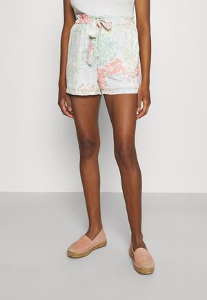 Woman wearing white floral high-waist paperbag shorts with tied belt and pink slip-on espadrille shoes standing against a plain white wall.