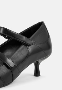 Chaussure à talon haut en cuir noir avec un bout arrondi, dotée d'une lanière sur le coup de pied et d'un talon élancé et effilé. Texture lisse.