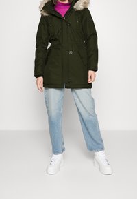 Parka vert foncé avec capuche en fausse fourrure, doté de poches avant, d'une taille cintrée et de fermetures à boutons. Associée à un jean large bleu clair et des baskets blanches.