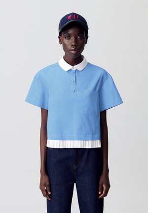 Chemise bleue à manches courtes, avec col blanc, poche avant et détail de bas plissé blanc, superposée sur un pantalon en denim foncé.