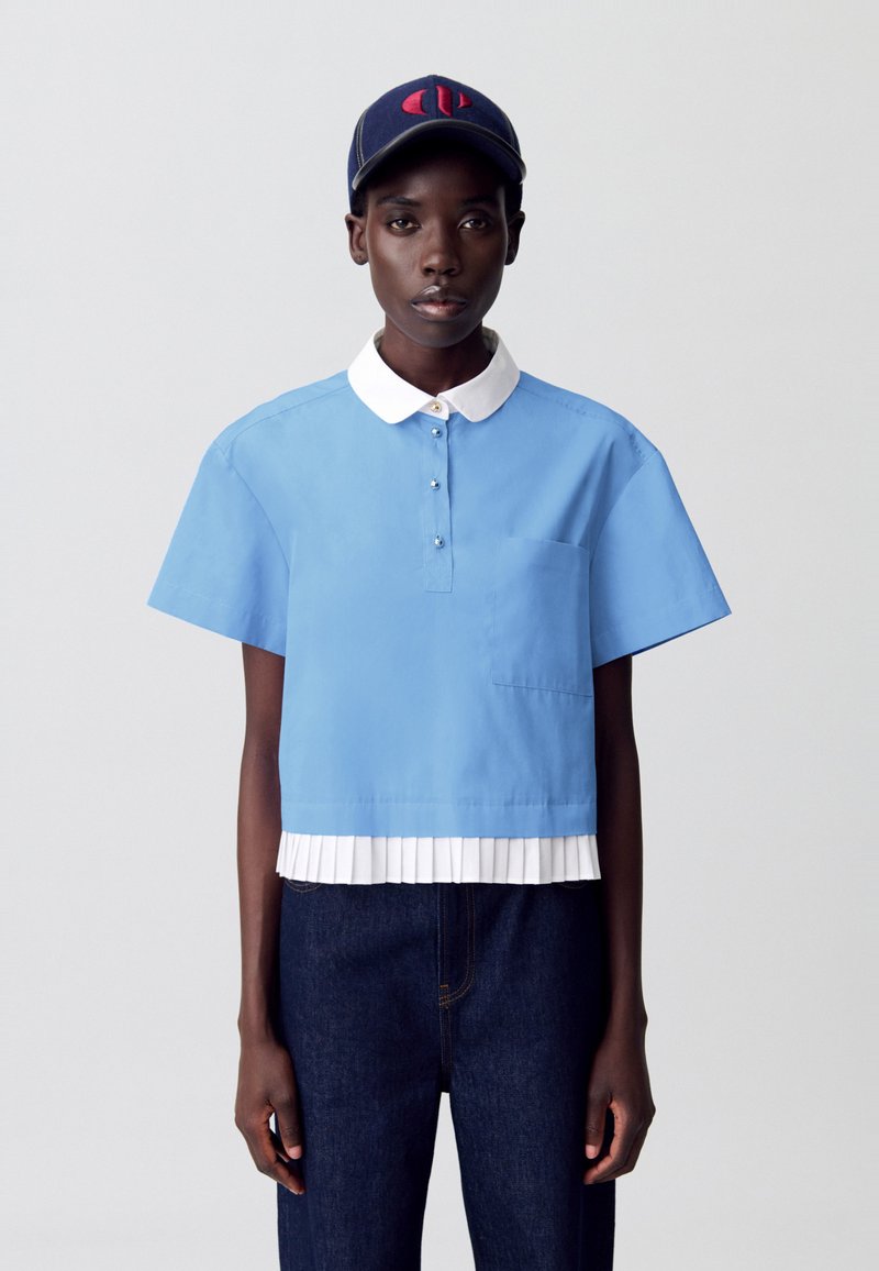 Camisa azul recortada de mangas cortas, con cuello blanco, bolsillo frontal y detalle de dobladillo blanco plisado, superpuesta sobre pantalones de mezclilla oscuros.