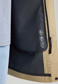 Beige windproof jacket with black inner lining, featuring waterproof ratings and taped seams. Zippered front and textured fabric.