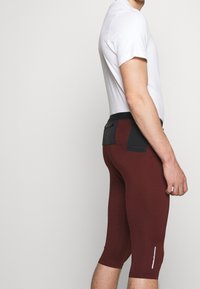 Pantalones cortos de ciclismo burdeos con un diseño ajustado y corte a la altura de la rodilla, que presentan una cinturilla negra y un acento reflectante. La camiseta es blanca.