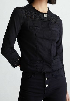 Frau trägt schwarze taillierte Jacke mit Ösenausschnitten an Schultern und Manschetten, am Kragen mit dekorativer silberner Brosche geschlossen.