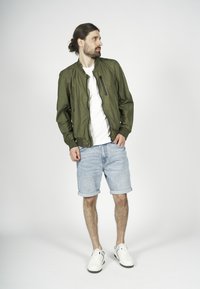 Giacca bomber verde oliva con tasca con zip, T-shirt bianca, pantaloni shorts in denim azzurro arrotolati e sneakers bianche con dettagli neri.