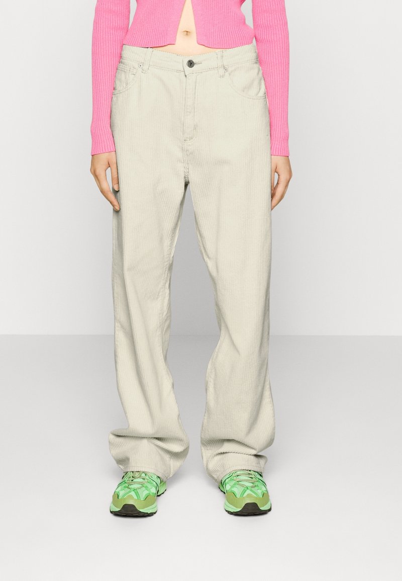 Personne portant un pantalon en velours côtelé beige ample, un pull court côtelé rose et des baskets vert néon, debout devant un fond uni.