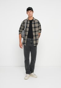 Camiseta negra debajo de una camisa de cuadros en tonos gris y verde, combinada con jeans negros y zapatillas blancas. Un bolso gris cuelga del hombro.