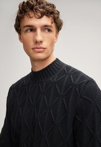 Schwarzer Strickpullover mit hohem Kragen, der ein geometrisches Muster und einen strukturierten Stoff aufweist. Nahaufnahme des Ausschnitts und der Schulter.