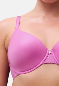 Frau trägt einen glatten, leuchtend pinkfarbenen Bügel-BH mit verstellbaren Trägern und einem kleinen dekorativen Anhänger in der Mitte vorne.