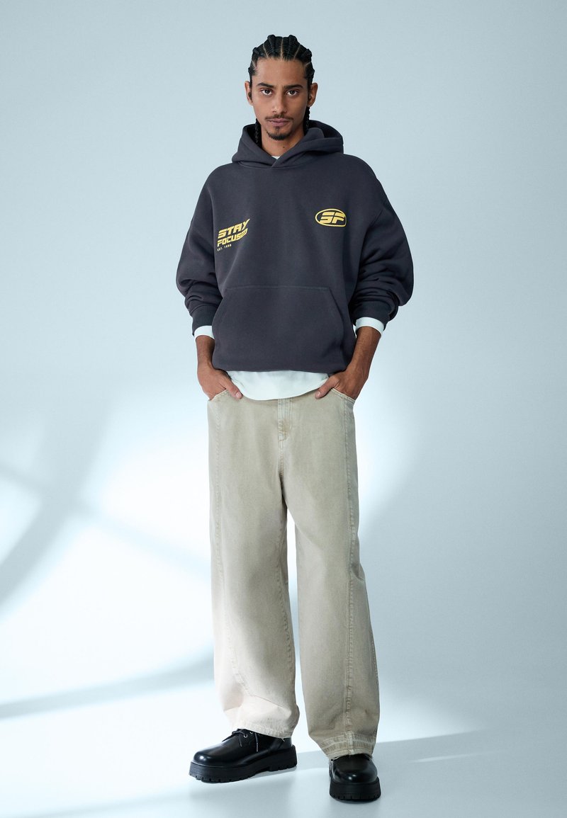 Mand med fletninger iført mørkegrå hættetrøje med gul tekst, beige vidde bukser og sorte sko, stående med hænderne i lommerne.