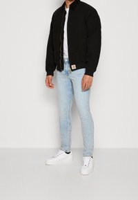 Hombre vestido con una chaqueta negra de cremallera, una camiseta blanca, jeans ajustados de color azul claro y zapatillas blancas, de pie contra un fondo blanco liso.