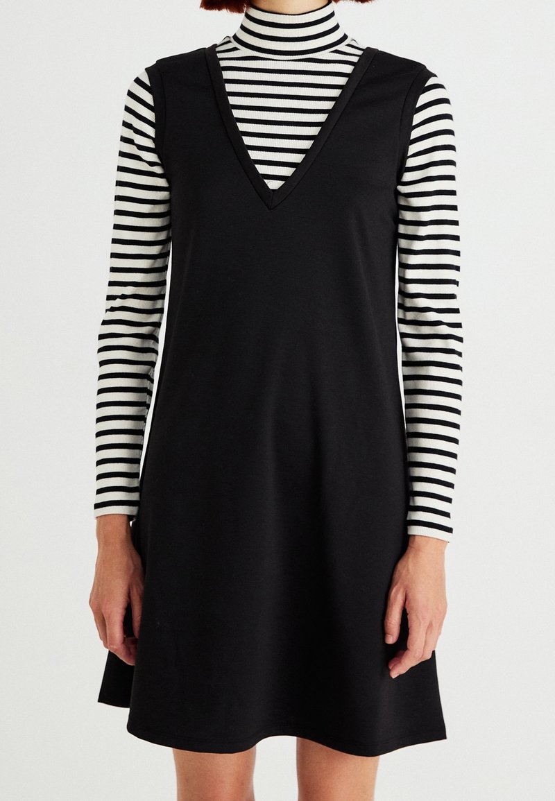 Femme portant une robe noire sans manches à col en V sur un haut à col roulé rayé noir et blanc à manches longues, debout devant un fond blanc.