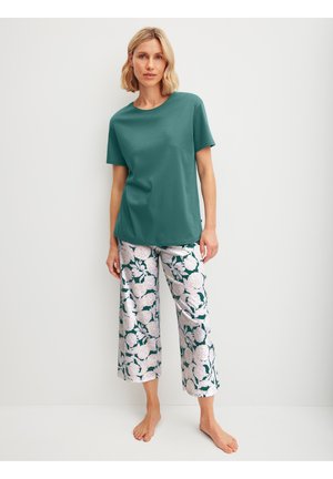 Femme debout pieds nus portant un t-shirt à manches courtes bleu sarcelle et un pantalon large à imprimé floral sur un fond blanc uni.